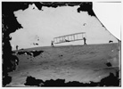  Start of a glide; Wilbur in motion at left holding one end of glider (rebuilt with single vertical rudder), Orville lying prone in machine, and Dan Tate at right; Kitty Hawk, North Carolina 

